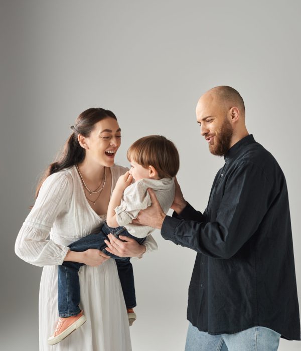 cheerful modern parents smiling and holding their little child on gray backdrop, family concept